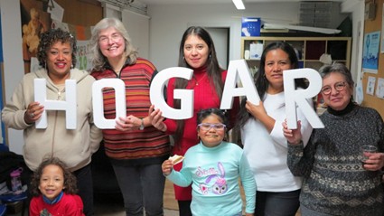 En la imagen aparece un grupo de seis mujeres y una niña pequeña en un espacio interior, sonriendo a cámara. Cuatro de las mujeres sostienen grandes letras blancas que forman la palabra “HOGAR”, mientras la niña, situada en el centro, lleva gafas y una sudadera azul claro y sostiene un trozo de comida, transmitiendo una sensación de convivencia, calidez y comunidad.